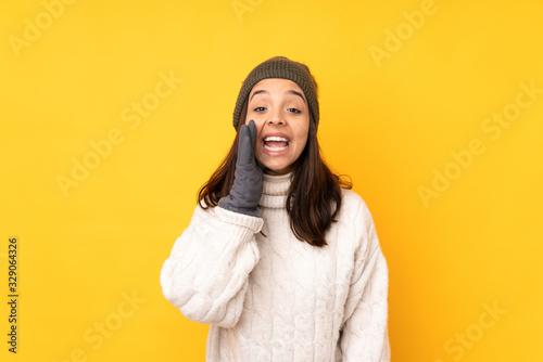 Young woman with winter hat over isolated yellow background shouting with mouth wide open