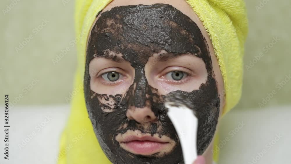 A girl with a towel on her head puts purifying black mask on her face by a cosmetic spatula. Charcoal face mask or black clay mud. Natural cosmetic concept. Skin care, acne treatment, cleansing skin. A girl with a towel on her head puts purifying black mask on her face by a cosmetic spatula. Charcoal face mask or black clay mud. Natural cosmetic concept. Skin care, acne treatment, cleansing skin.