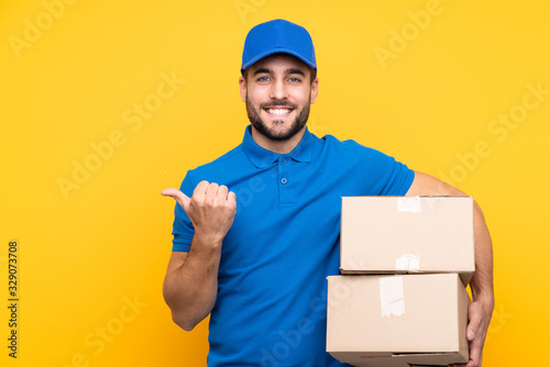 Delivery man over isolated yellow background pointing to the side to present a product