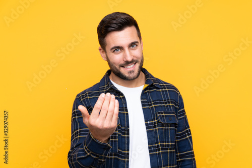 Young handsome man with beard over isolated yellow background inviting to come with hand. Happy that you came