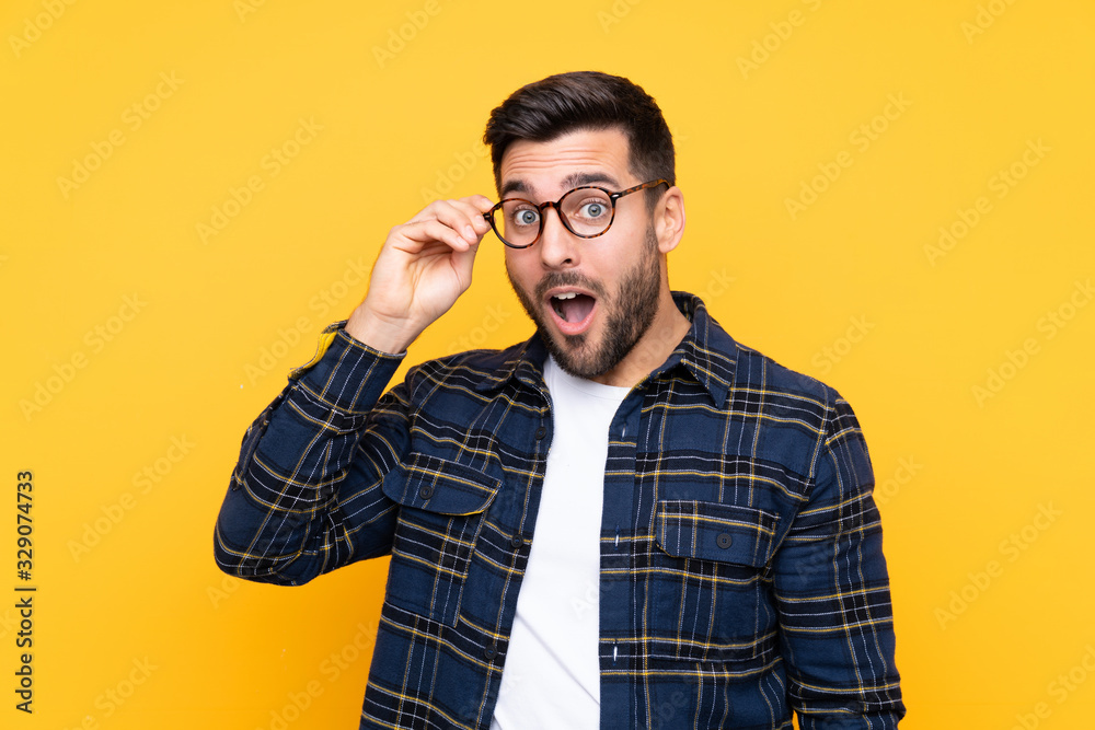 Young handsome man with beard over isolated yellow background with glasses and surprised Young handsome man with beard over isolated yellow background with glasses and surprised