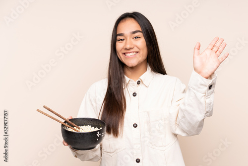 Young Indian woman isolated on beige background saluting with hand with happy expression while holding a bowl of noodles with chopsticks