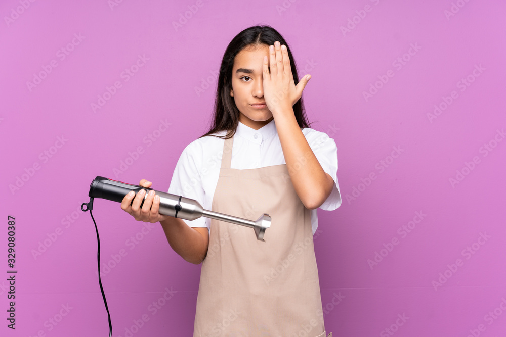 Indian woman using hand blender isolated on purple background covering a eye by hand Indian woman using hand blender isolated on purple background covering a eye by hand