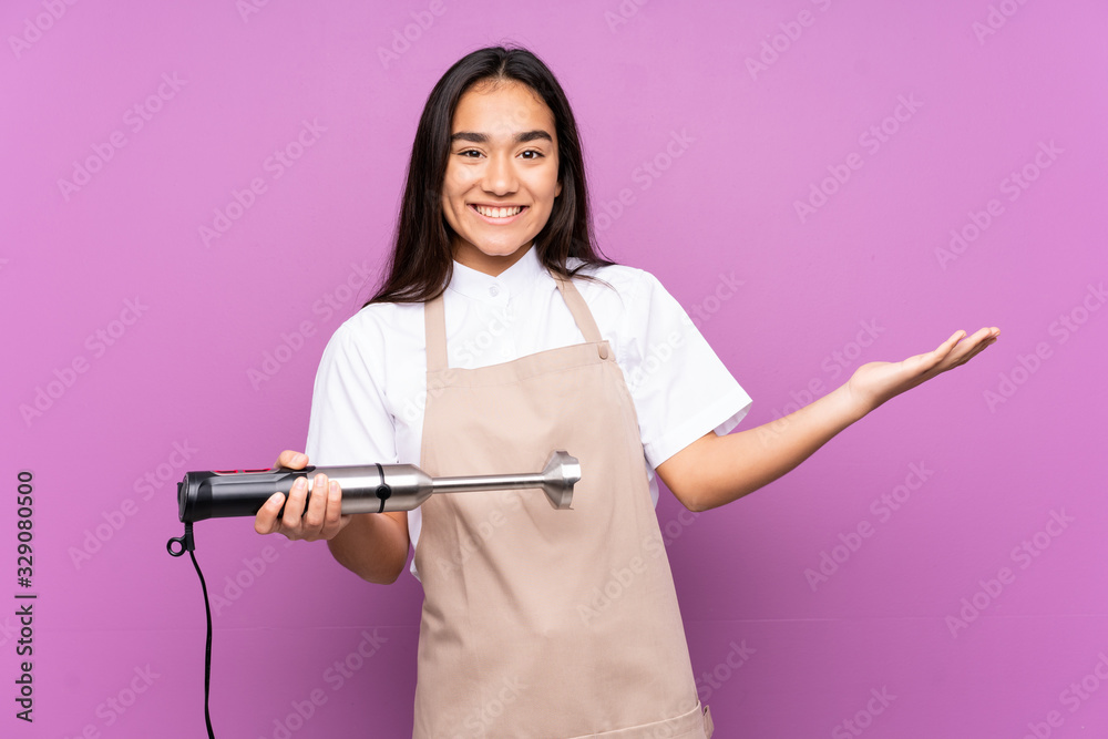 Indian woman using hand blender isolated on purple background extending hands to the side for inviting to come Indian woman using hand blender isolated on purple background extending hands to the side for inviting to come