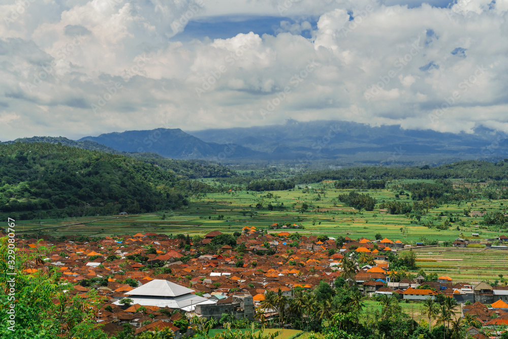 View of the valley with a traditional Balinese village. Orange roofs of houses, coconut palms and rice fields against the background of mountains and clouds.