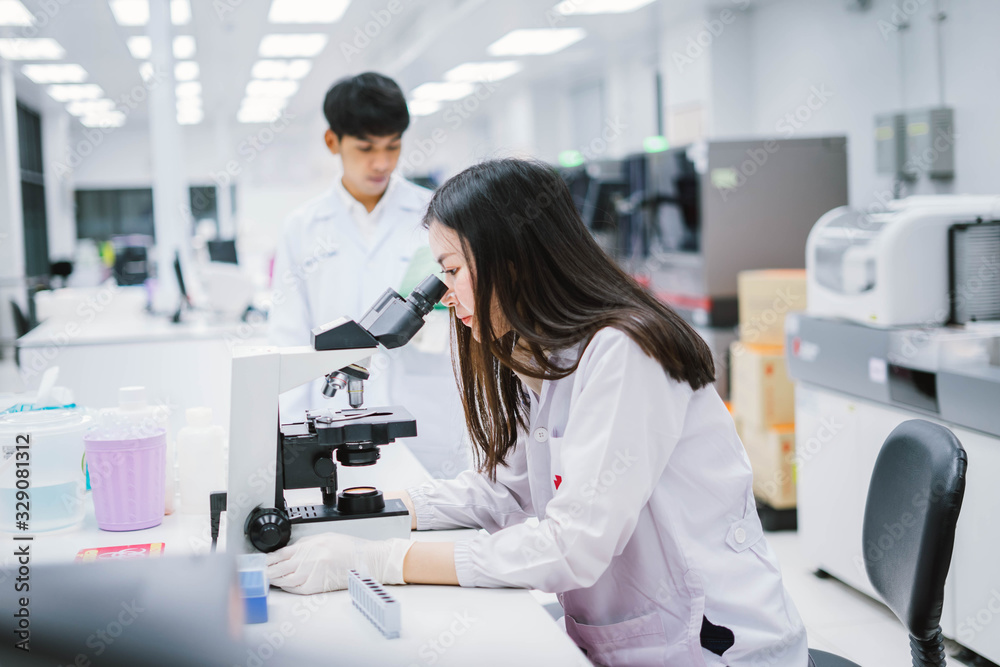 Two medical scientist working in Medical laboratory , young female scientist looking at microscope and young male look at report on his hand. select focus on female scientist. Two medical scientist working in Medical laboratory , young female scientist looking at microscope and young male look at report on his hand. select focus on female scientist.