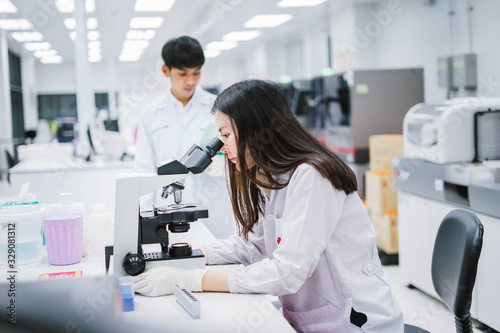 Two medical scientist working in Medical laboratory , young female scientist looking at microscope and young male look at report on his hand. select focus on female scientist.