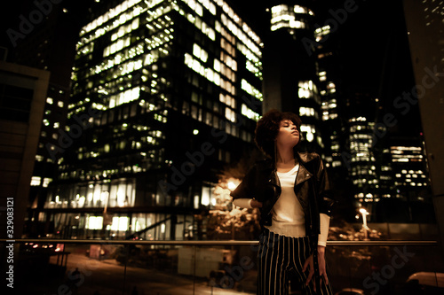Stylish african american woman in night city. Woman standing with hand on waist with eyes closed. Blurred high-rise building with yellow light on background