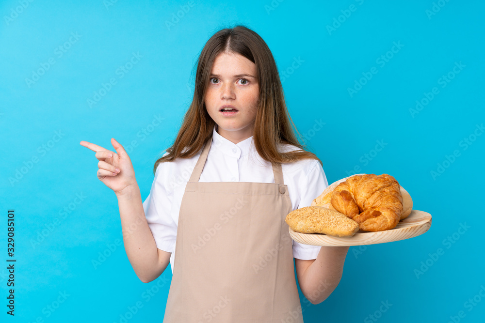 Ukrainian teenager chef uniform. Female baker holding a table with several breads surprised and pointing side Ukrainian teenager chef uniform. Female baker holding a table with several breads surprised and pointing side