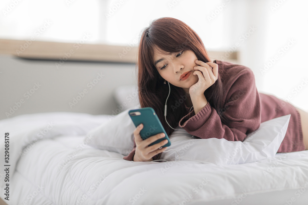 Asian happy woman sitting on bed is enjoying listening to music and watching entertainment from her smartphone with earphones. Asian happy woman sitting on bed is enjoying listening to music and watching entertainment from her smartphone with earphones.