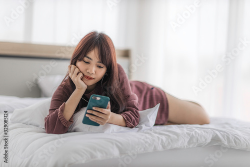 Asian happy woman sitting on bed is enjoying listening to music and watching entertainment from her smartphone with earphones.