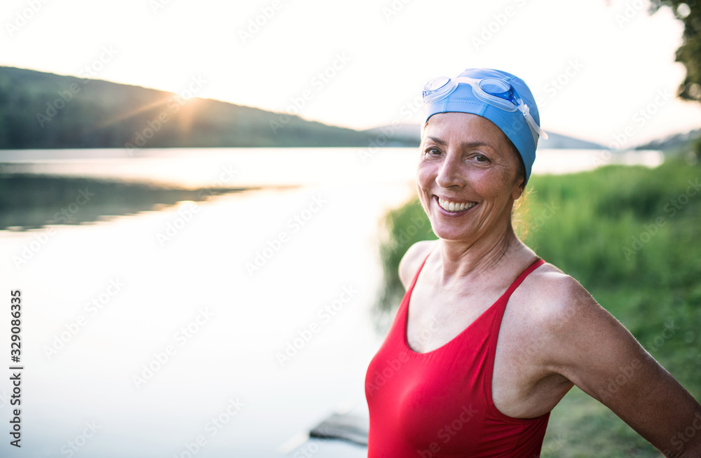 Senior woman in swimsuit standing by lake outdoors before swimming.