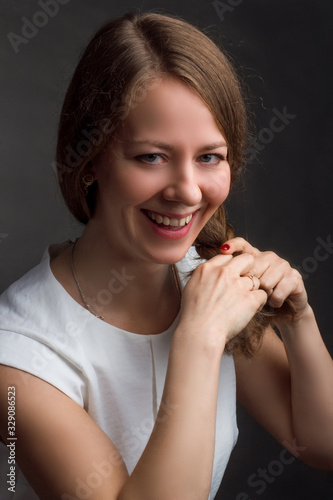 A photo of a woman with brown straight hair posing in white shirt on dark background in Studio. laughing, plaits her hair.