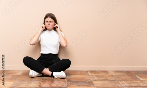 Ukrainian teenager girl sitting on the floor frustrated and covering ears