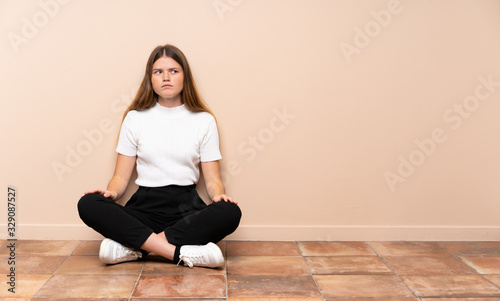 Ukrainian teenager girl sitting on the floor making doubts gesture looking side