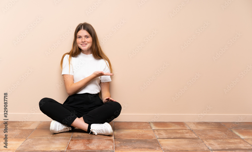 Ukrainian teenager girl sitting on the floor presenting an idea while looking smiling towards Ukrainian teenager girl sitting on the floor presenting an idea while looking smiling towards