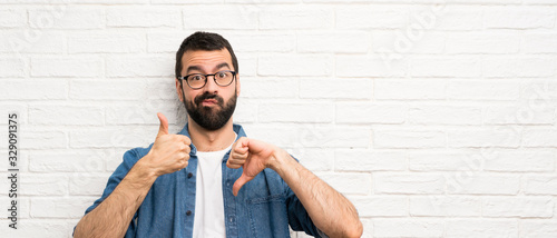 Photos Handsome man with beard over white brick wall making good-bad sign