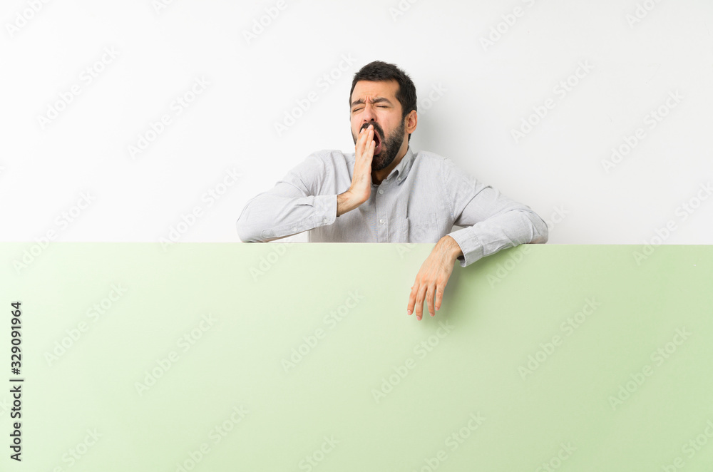 Young handsome man with beard holding a big green empty placard yawning and covering wide open mouth with hand Young handsome man with beard holding a big green empty placard yawning and covering wide open mouth with hand