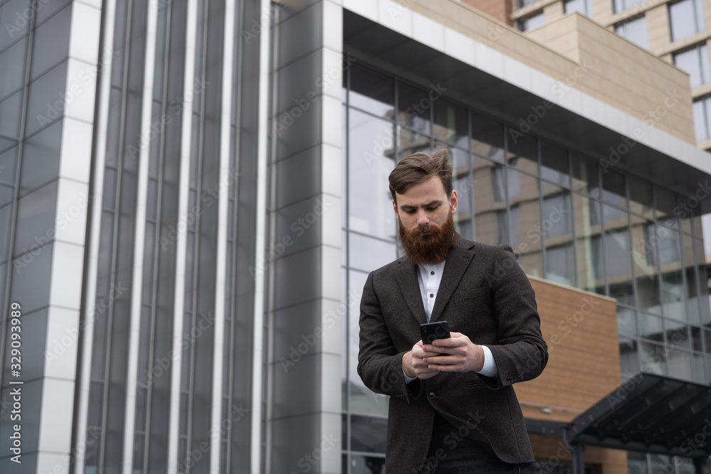 Young bearded Businessman holding mobile smartphone using app texting sms message wearing jacket outdoor. Successful entrepreneur dressed in formal wear communicating with a business partner online.