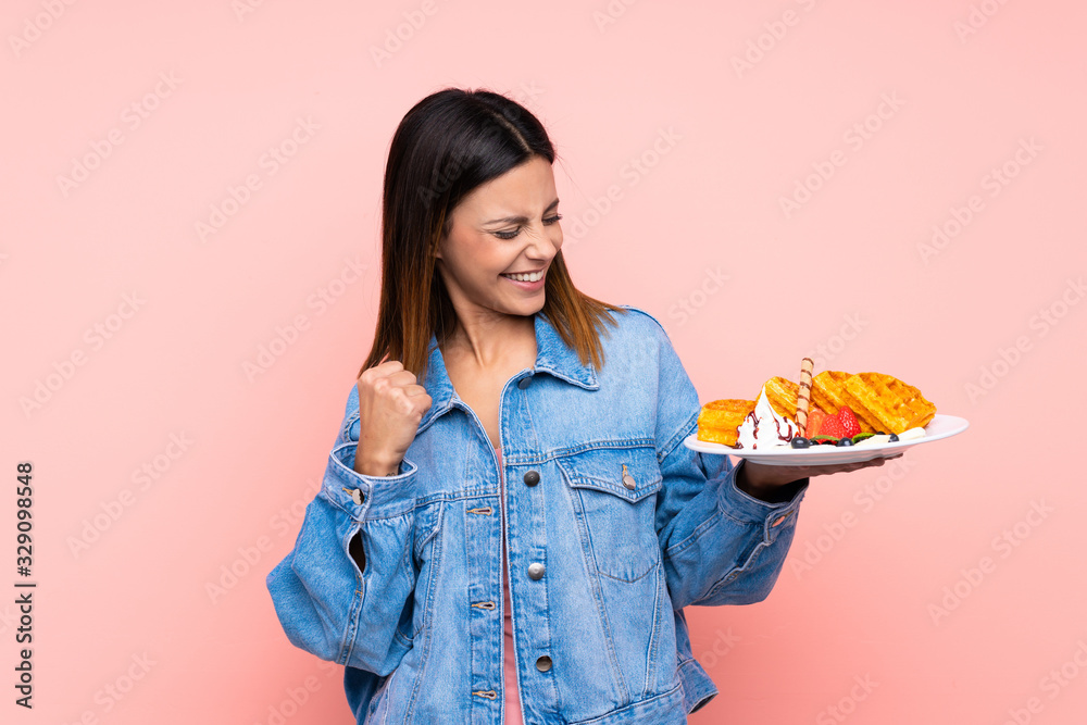 Brunette woman holding waffles over isolated pink background celebrating a victory