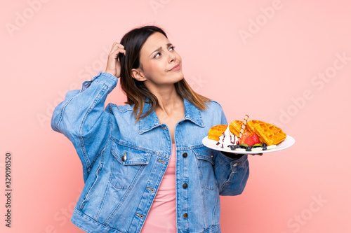 Brunette woman holding waffles over isolated pink background having doubts and with confuse face expression
