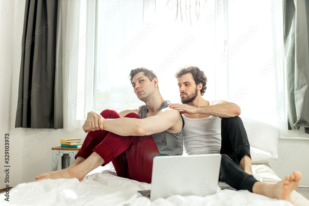 young caucasian gay couple in quarrel at home, offended man sit waiting for apologies from his same-sex partner. LGBT and alternative relationship concept