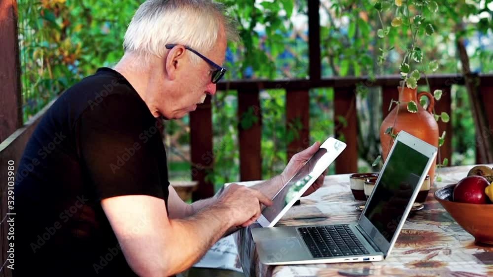 Senior man using laptop and tablet in the porch garden, slow motion Senior man using laptop and tablet in the porch garden, slow motion