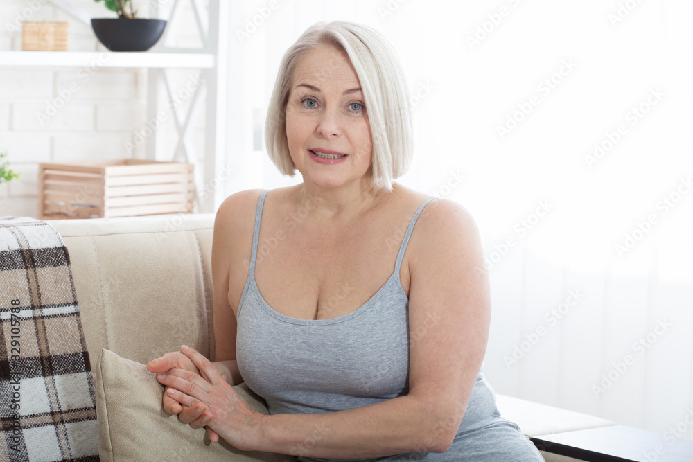 Active beautiful middle-aged woman smiling friendly and looking in camera in living room. Woman's face close up. Realistic images without retouching with their own imperfections. Selective focus. Active beautiful middle-aged woman smiling friendly and looking in camera in living room. Woman's face close up. Realistic images without retouching with their own imperfections. Selective focus.