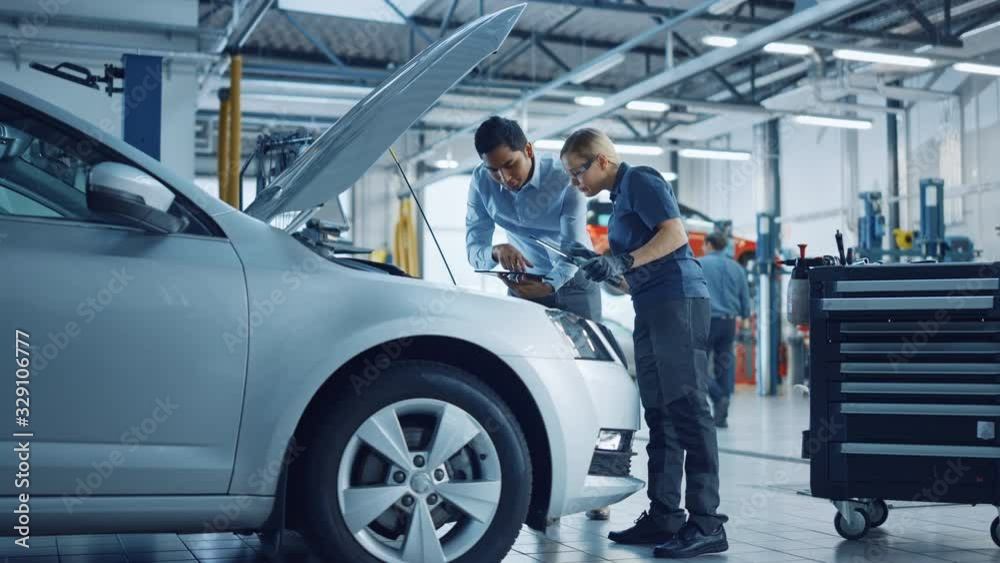 Manager Checks Tasts on a Tablet Computer and Explains an Engine Breakdown to an Female Mechanic. Car Service Employees Inspect Car's Engine Bay with a LED Lamp. Modern Clean Workshop.