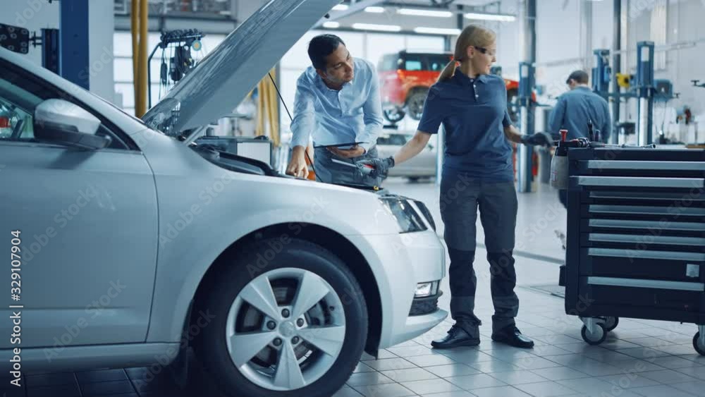 Manager Checks Diagnostics Results on a Tablet Computer and Explains an Engine Breakdown to an Female Mechanic. Car Service Employees Inspect Car's Engine Bay with a LED Lamp. Modern Clean Workshop.