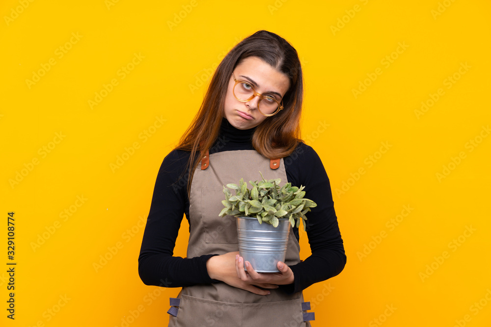 Young gardener girl holding a plant over isolated yellow background sad Young gardener girl holding a plant over isolated yellow background sad