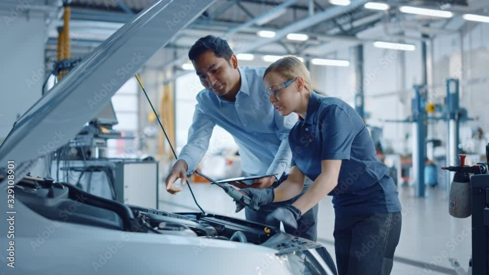 Instructor with a Tablet Computer is Giving a Task for a Future Mechanic. Female Student Inspects the Car Engine. Assistant is Checking the Cause of a Breakdown in the Vehicle in a Car Service. Instructor with a Tablet Computer is Giving a Task for a Future Mechanic. Female Student Inspects the Car Engine. Assistant is Checking the Cause of a Breakdown in the Vehicle in a Car Service.