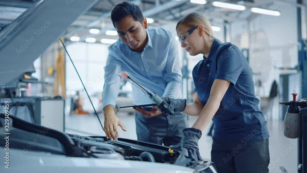 Instructor with a Tablet Computer is Giving a Task for a Future Mechanic. Female Student Inspects the Car Engine. Assistant is Checking the Cause of a Breakdown in the Vehicle in a Car Service. Instructor with a Tablet Computer is Giving a Task for a Future Mechanic. Female Student Inspects the Car Engine. Assistant is Checking the Cause of a Breakdown in the Vehicle in a Car Service.