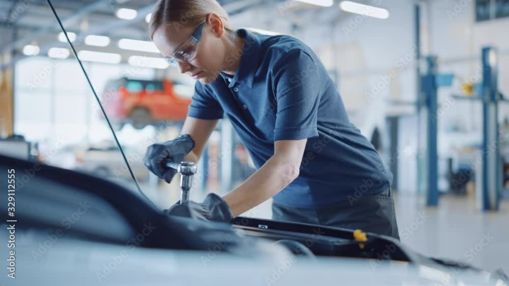 Beautiful Empowering Female Mechanic is Working on a Car in a Car Service. Woman in Safety Glasses is Working on an Usual Car Maintenance. She's Using a Ratchet. Modern Clean Workshop with Cars. Beautiful Empowering Female Mechanic is Working on a Car in a Car Service. Woman in Safety Glasses is Working on an Usual Car Maintenance. She's Using a Ratchet. Modern Clean Workshop with Cars.