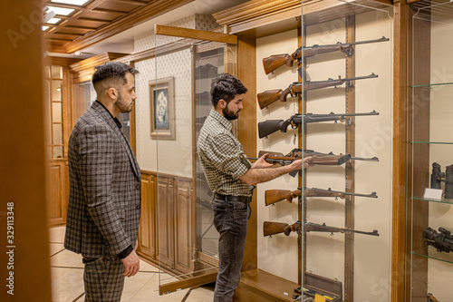 young caucasian men discuss weapon in store. handsome confident offer the best rifle in their collection, in their showcase. customer want to buy it © Roman