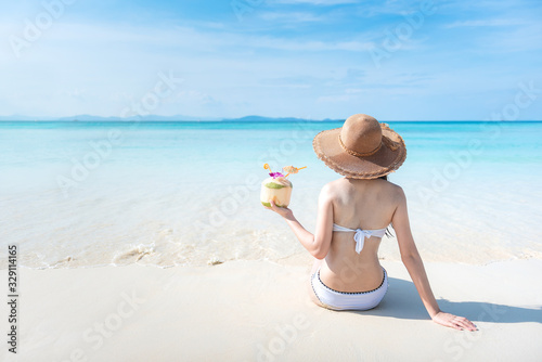 beautiful young tan skin asian woman sitting on the beach wearing bikini ,beach hat with hand holding the coconut drink. relaxing summer holiday with sunny blue sky on vacation concept