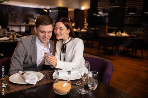 attractive caucasian lady show something interesting on smartphone to boyfriend. guy look at screen of mobile phone and smile, laugh. happy couple in restaurant