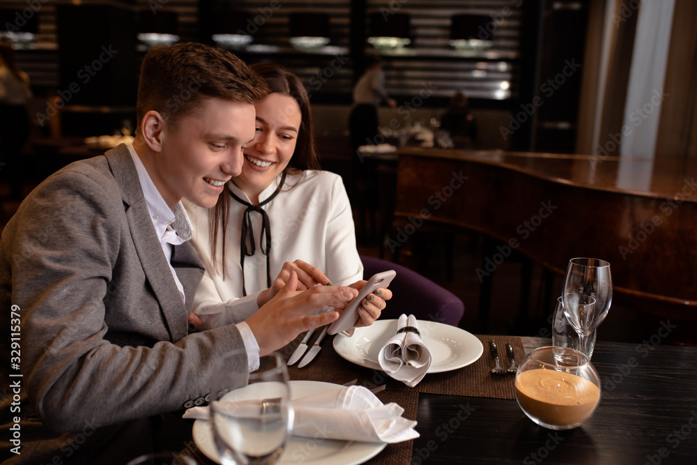 young caucasian couple have dinner together, look at smartphone in restaurant. man show cool video to girlfriend. indoors. man and woman enjoy spending time together