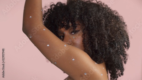 Mixed race black woman with curly hair covered by crystal makeup on pink background in studo dancing lifted her hand to cover her face photo