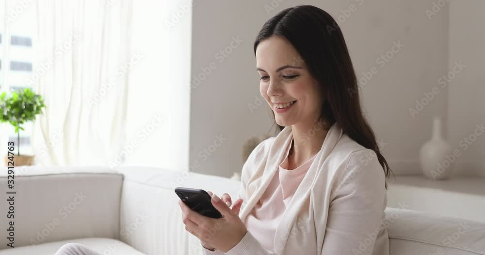 Happy millennial woman checking social media feed on smartphone at home. Smiling girl having fun spending time at home with mobile gadget. Relaxed lady using phone apps playing games relaxing on sofa Happy millennial woman checking social media feed on smartphone at home. Smiling girl having fun spending time at home with mobile gadget. Relaxed lady using phone apps playing games relaxing on sofa