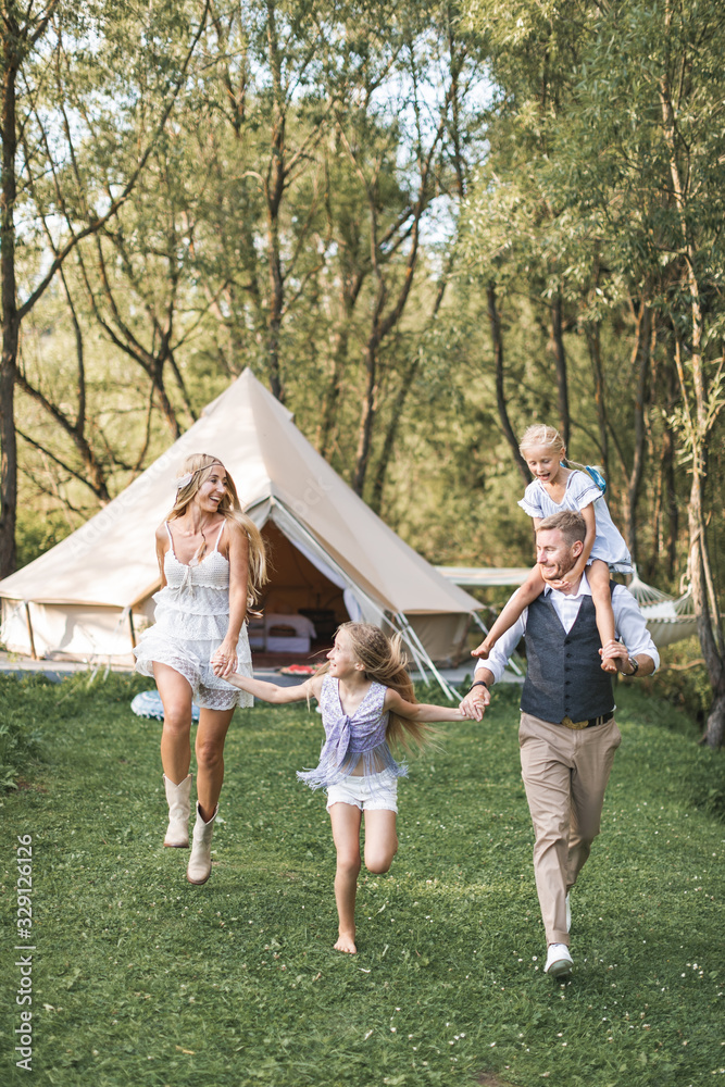 Fototapeta premium Happy Caucasian family, father mother and two little daughters, having fun outdoors in park or camping, enjoying time and laughing together, holding hands and running. Love and parenthood concept