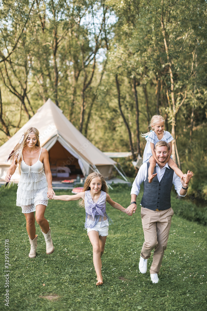 Fototapeta premium Young family in boho casual wear, dad, mom and two daughters holding hands and running and jumping in a green park, big wigwam tent on the background. Family walk, outdoors