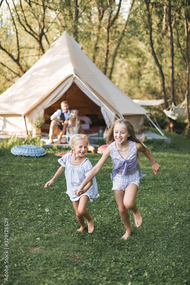 Fototapeta premium Beautiful young family spending time together in summer park or meadow, having a picnic. Young parents sitting near wigwam, while two little girls daughters running and laughing
