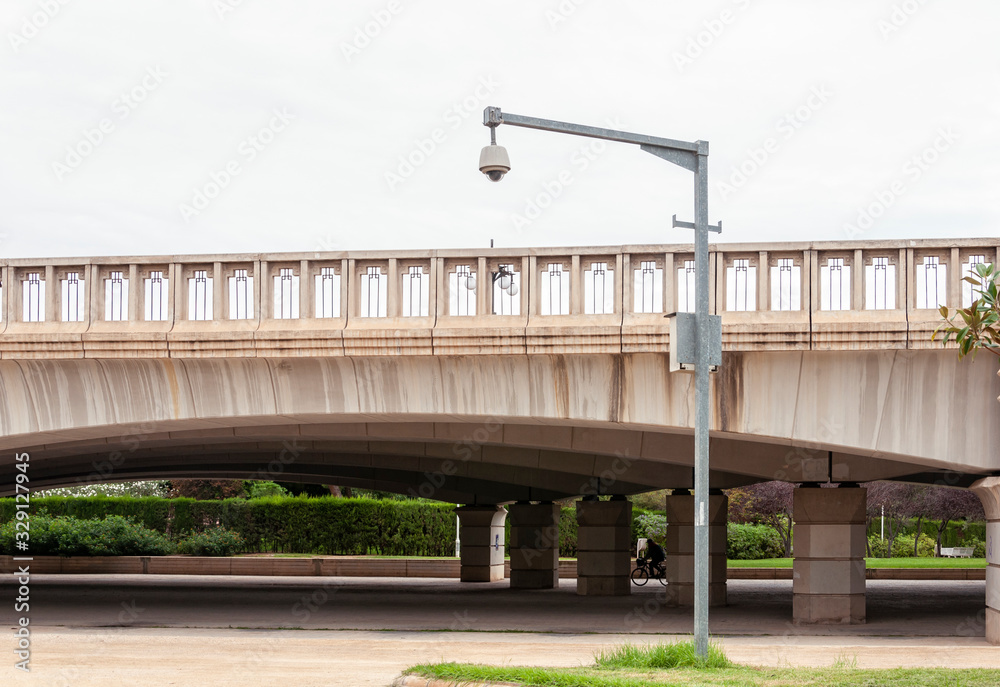 Surveillance camera on a metal pole, next to a bridge over a park 