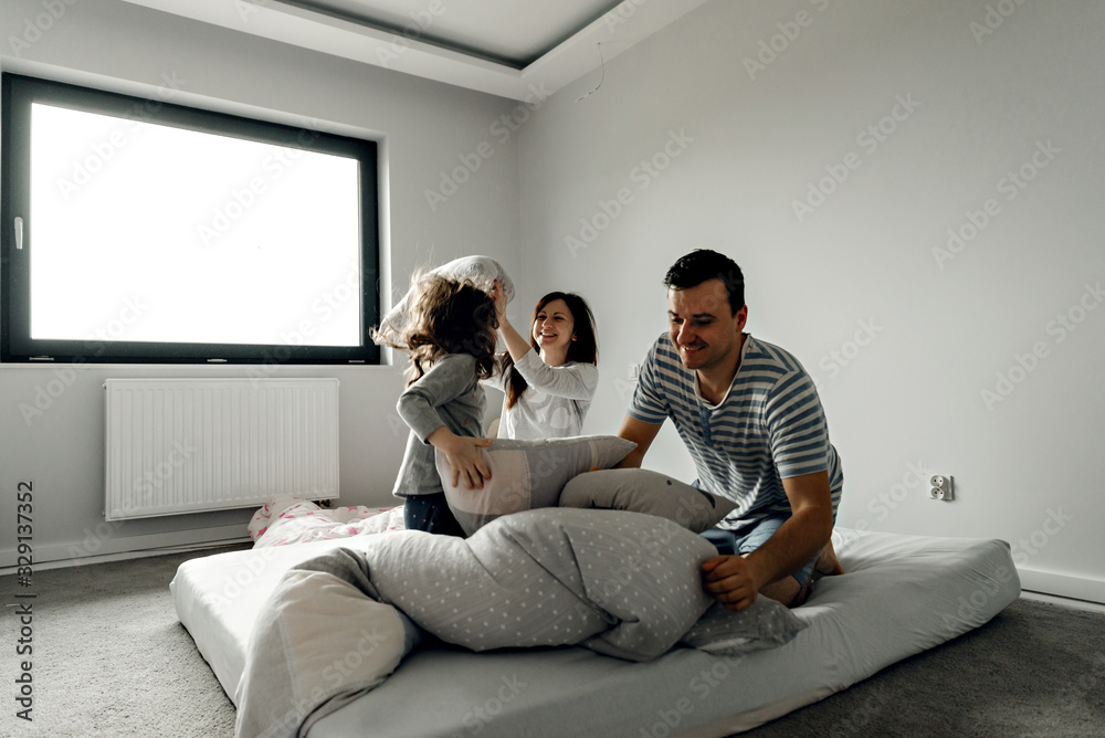 Naklejka premium people, family and morning concept - happy child with parents having pillow fight in bed at home,family with a funny pillow fight on the bed. Parents spend their free time with their daughters.