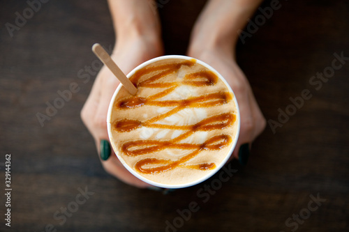 Caramel Big Coffee paper cup with milk in womans hand on the wooden table. cappuccino or latte drink, cup of coffee on table flat lay view. Cup of cafe au lait. Milk paintings. Hot Coffee for girl
