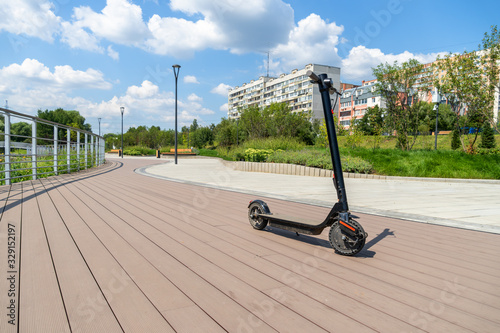 An electric black scooter stands on the bandwagon on the street. City park with wooden flooring along the promenade with railings. Sunny summer day. Modern city transport.