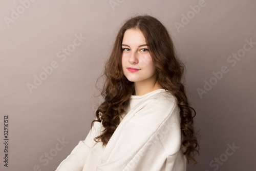 Beautiful young brunette teenager girl with wavy long curly thick hair in a white sweater on a gray background. Waist photo