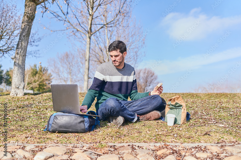 young man sitting on a park lawn eating a sandwich while working on his laptop