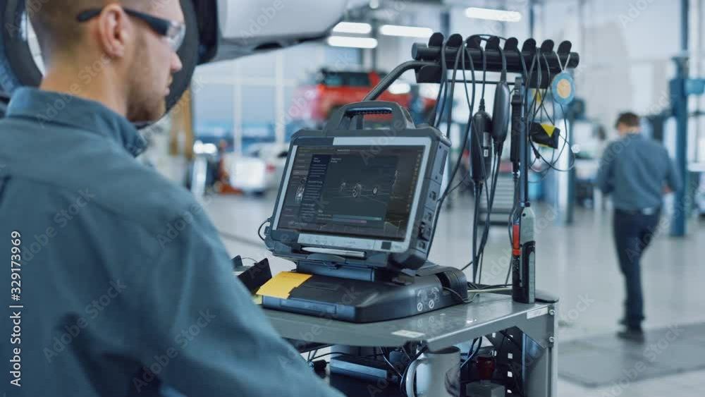 Car Service Manager or Mechanic is Running an Interactive Diagnostics Software on an Advanced Computer. Specialist Inspecting the Vehicle in Order to Find Broken Components and Errors in Data Logs. Car Service Manager or Mechanic is Running an Interactive Diagnostics Software on an Advanced Computer. Specialist Inspecting the Vehicle in Order to Find Broken Components and Errors in Data Logs.